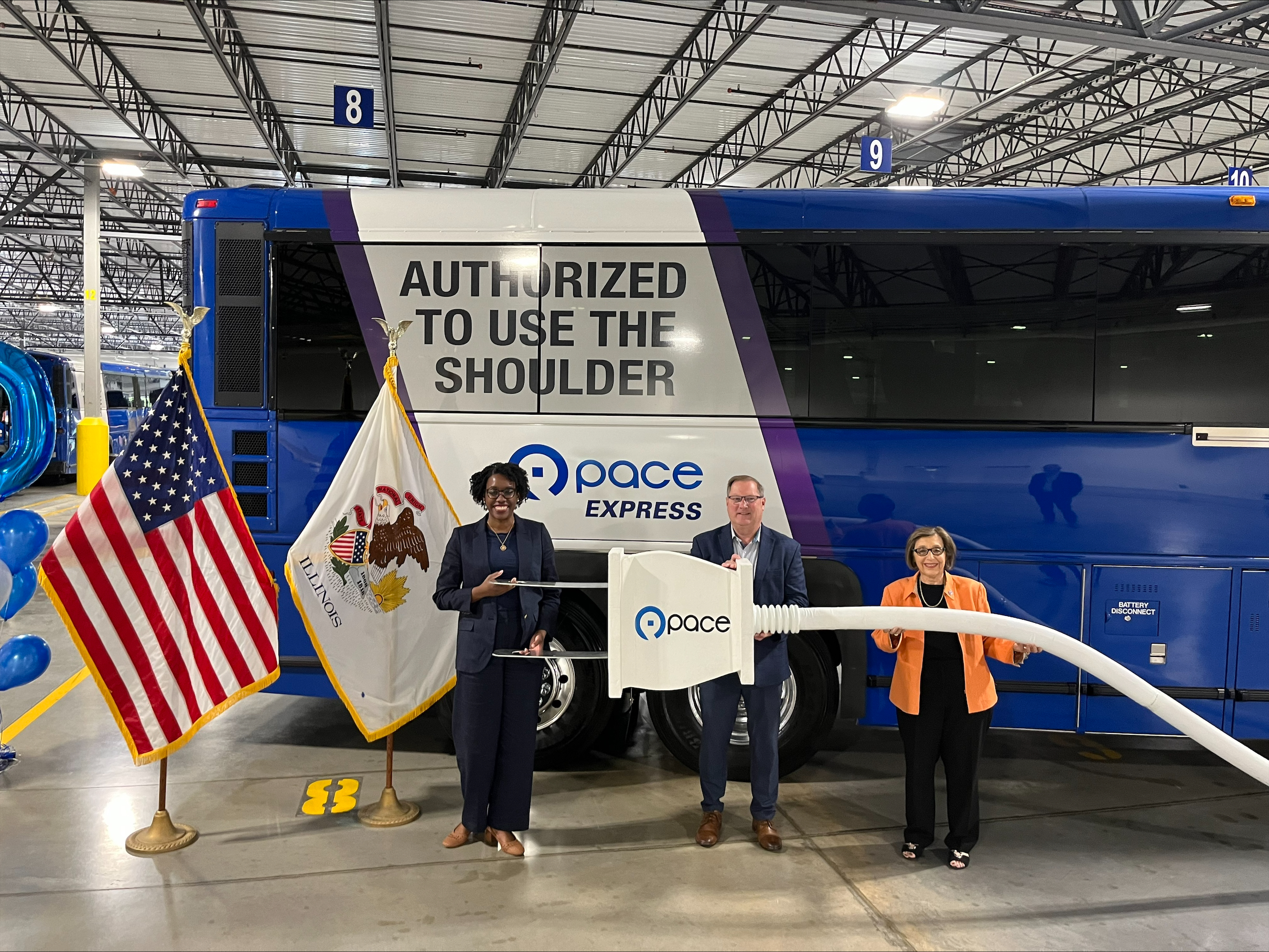 (L-R) U.S. Congresswoman Lauren Underwood, Pace Chairman Rick Kwasneski. and Pace Executive Director Melinda Metzger. in front of a Pace coach bus holding a big electric plug prop. 